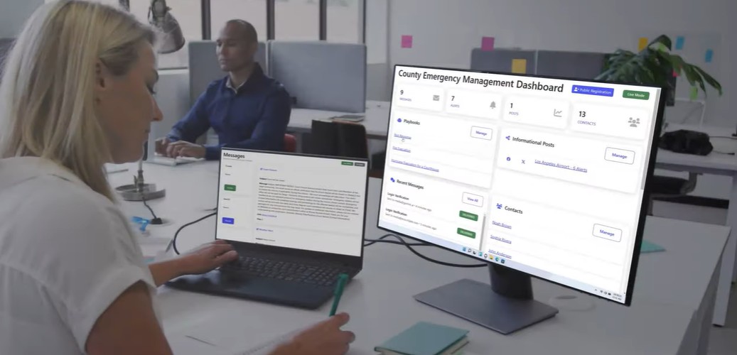 A woman works at a desk using a laptop and an external monitor displaying a “County Emergency Management Dashboard.” The screen shows alerts, messages, and contact management tools, suggesting real-time communication and coordination in a government office setting.